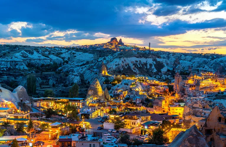 Panorámica nocturna de Göreme en Capadocia, Turquía, con hoteles cueva iluminados, chimeneas de hadas y el castillo de Uchisar al fondo bajo un cielo de atardecer.