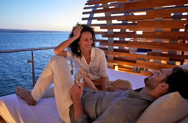 Pareja disfrutando de una velada al atardecer en una cabaña privada del Celebrity Flora, con copas de champagne y vistas al océano en las Islas Galápagos.