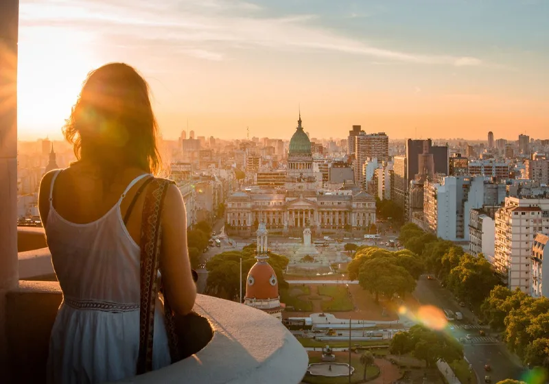 Una mujer joven con un vestido blanco contempla desde las alturas el Palacio del Congreso de la Nación en Buenos Aires, Argentina, durante un atardecer dorado que ilumina el paisaje urbano.