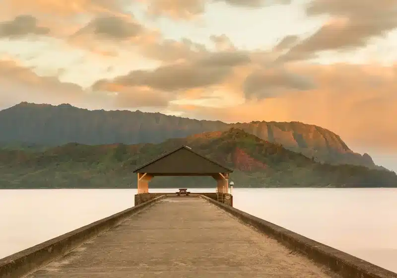 Perspectiva hacia el muelle cubierto de Hanalei Pier en la costa de Kauai, Hawái, con imponentes montañas verdes al fondo bajo un cielo nublado iluminado por el atardecer.