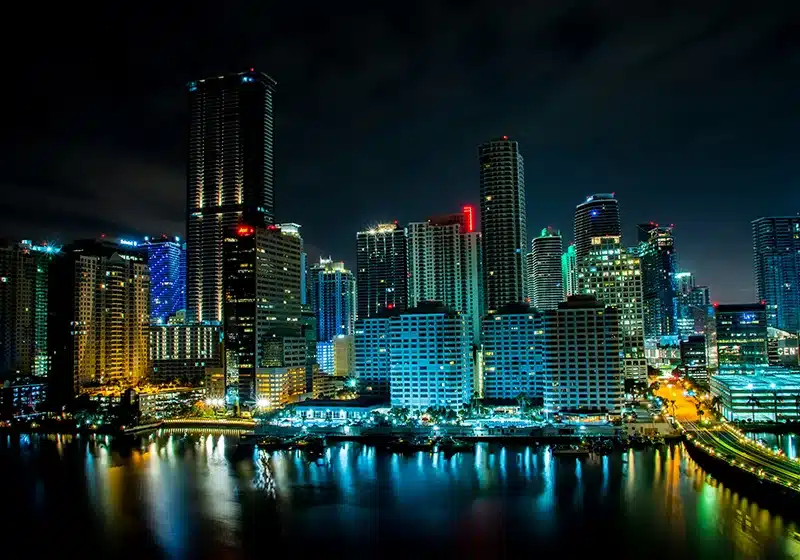 Vista nocturna del horizonte urbano del centro de Miami con rascacielos iluminados en tonos azules, verdes y dorados, reflejándose sobre el agua de la bahía y un puente con tráfico a la derecha.