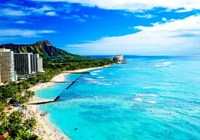 Vista aérea de la playa de Waikiki en Oahu, Hawái, mostrando aguas turquesas, hoteles en la costa y el icónico cráter Diamond Head al fondo bajo un cielo azul con nubes blancas.